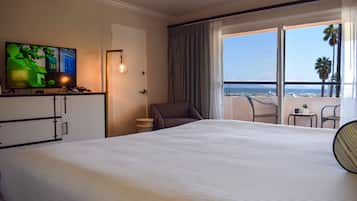 Habitación de hotel con una cama grande, un televisor sobre un soporte, una silla, una mesita y vistas a la playa a través de un balcón.