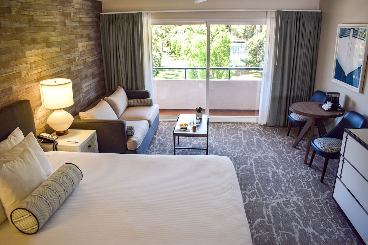 A hotel room with a large bed, a desk with a lamp, a sofa, a dining table with chairs, and a balcony with curtains.