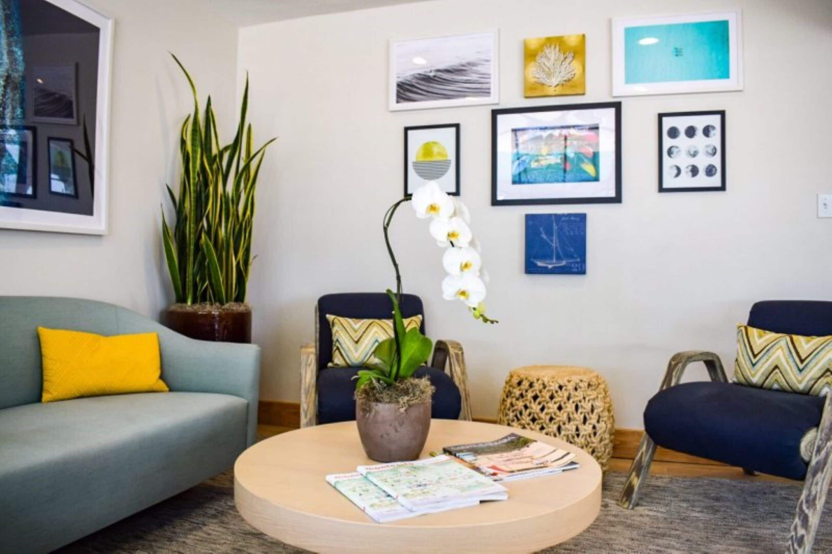 Un salon moderne avec une table basse ronde, un canapé et deux fauteuils. Des œuvres d’art encadrées ornent le mur, et une plante en pot est posée sur le sol.