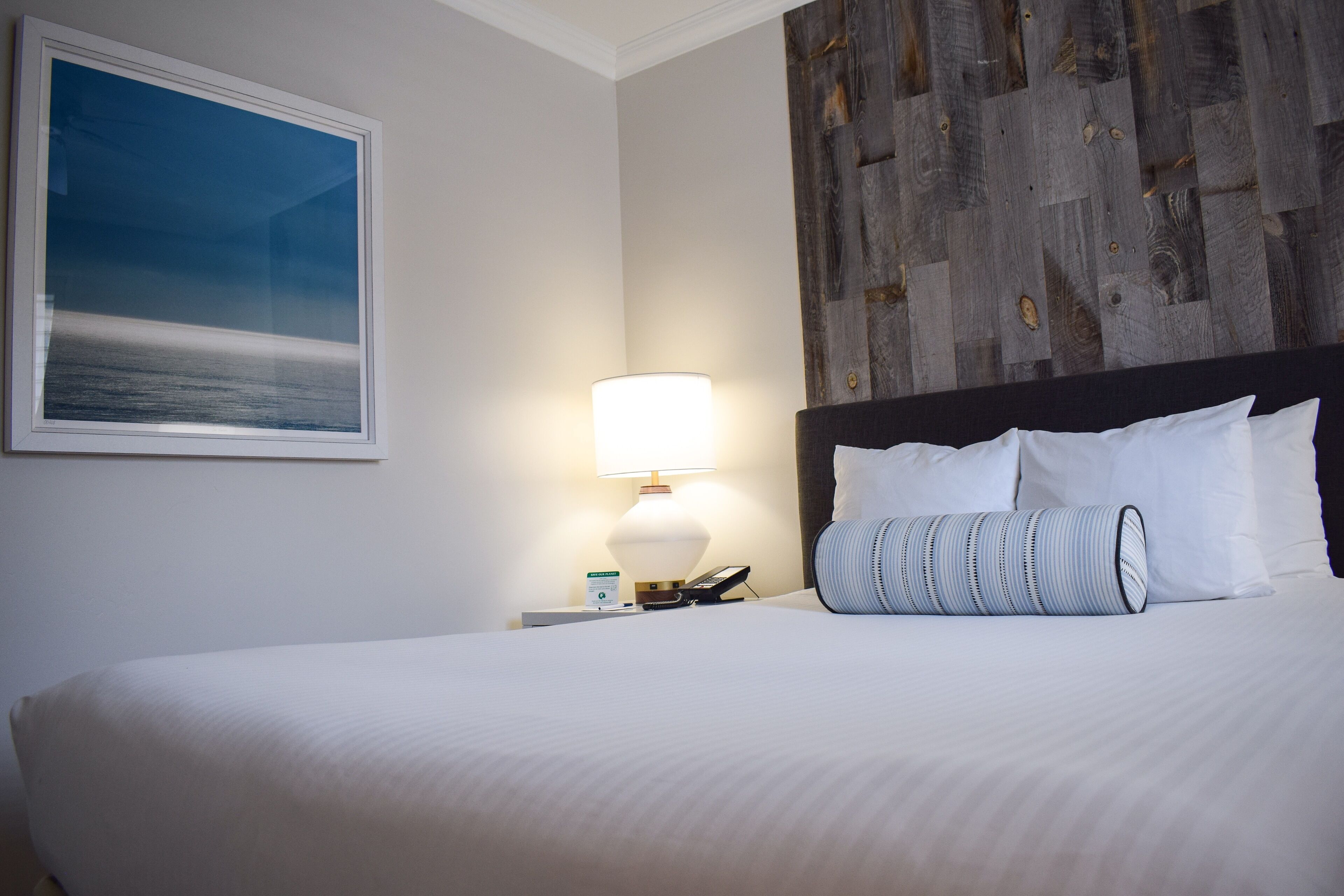 A neatly made bed with white linens, a bedside lamp, a phone, and a framed picture of a beach on the wall.