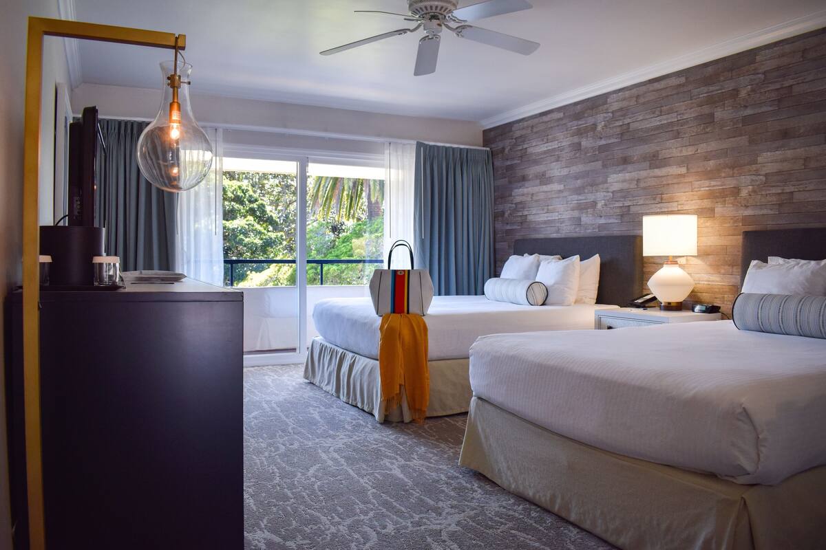 A hotel room with two beds, a ceiling fan, a large window with curtains, a wooden accent wall, and a desk with a lamp.