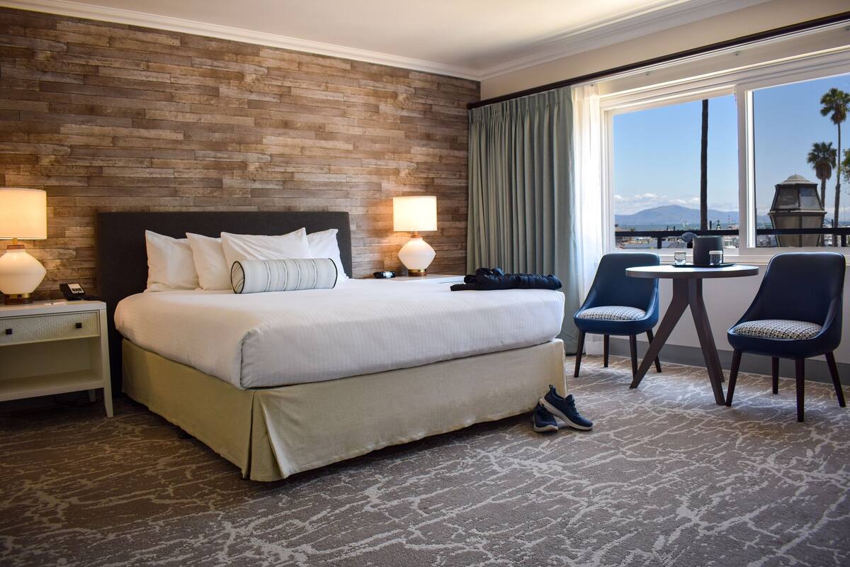 A hotel room with a large bed, two bedside tables with lamps, a small round table with two chairs, and a view of the outdoors through a large window.