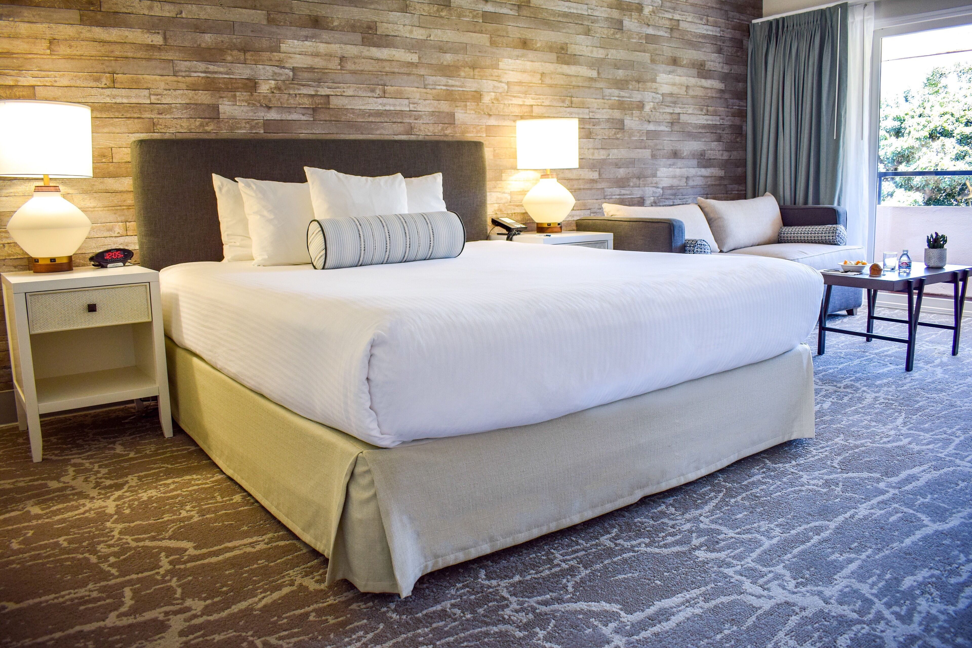 A large bed with white linens, two bedside tables with lamps, a small table with a chair, and a brick-patterned wall.