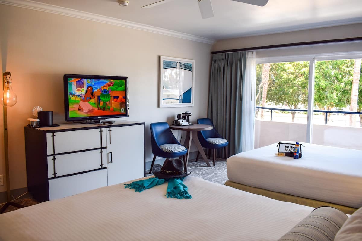 A hotel room with two beds, a television, a desk with a computer, and a view of trees through the window.