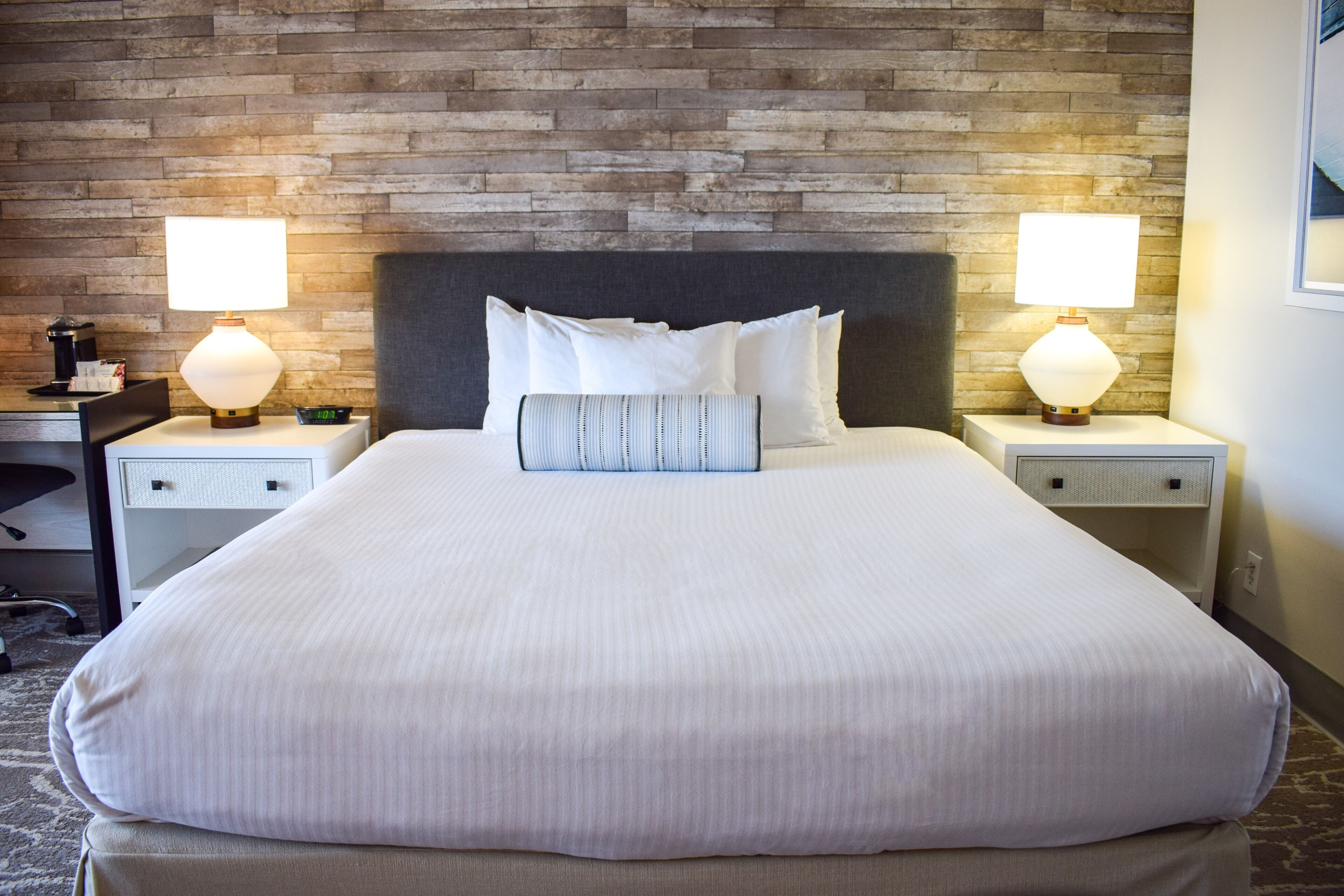 Een netjes opgemaakt bed met witte lakens, twee bedlampjes en een muur met baksteenmotief.