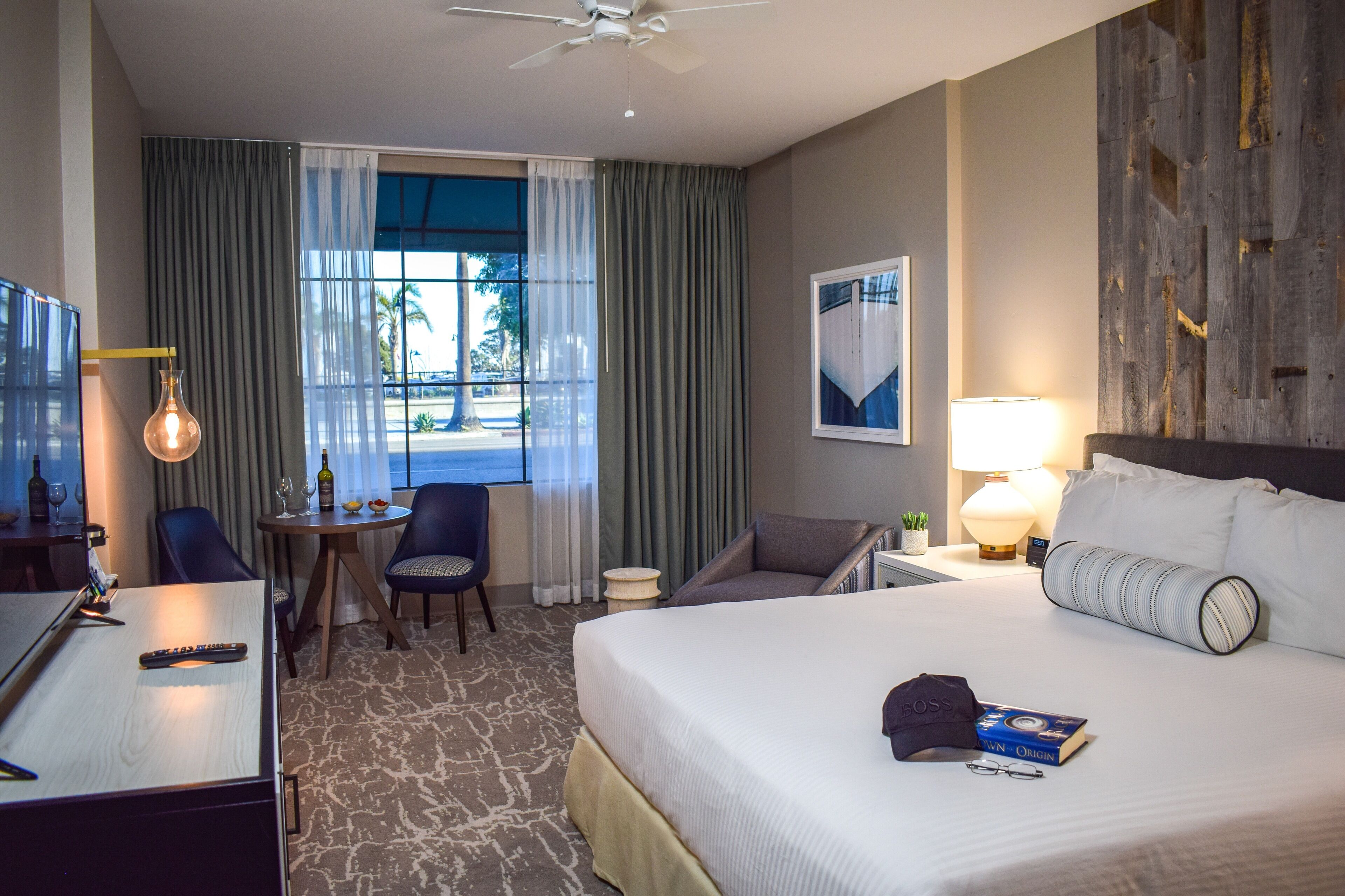 Quarto de hotel com cama, escrivaninha com controle remoto, cadeira, mesa pequena com garrafa e copos, luminária e vista para o exterior pela janela.