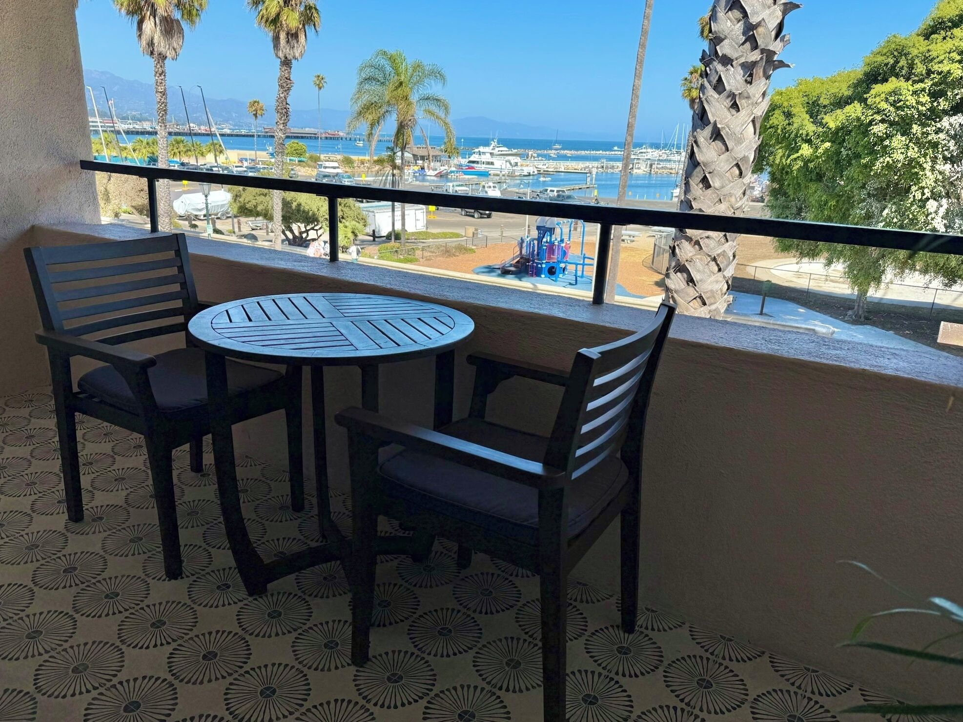 Un balcón con mesa y sillas, con vista a un puerto deportivo con barcos y palmeras.