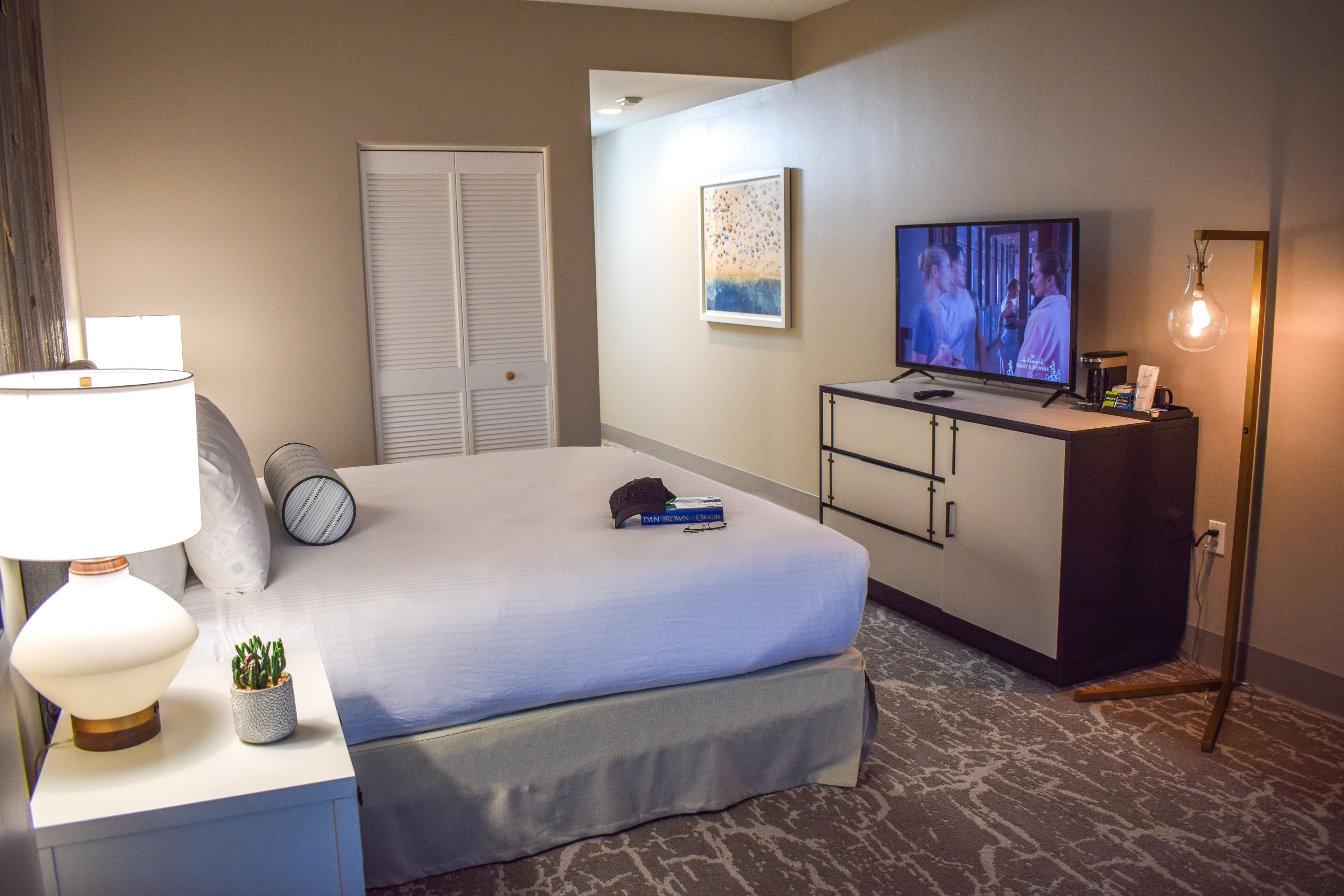 Een hotelkamer met een bed, een bureau met lamp, een televisie, een kast en een aan de muur hangend schilderij.