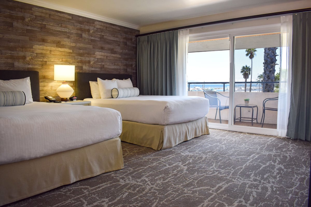 A hotel room with two beds, a nightstand, a lamp, and a balcony with a view of the beach.