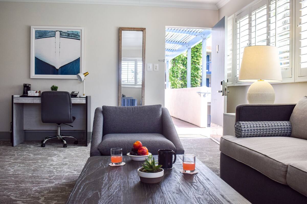 A modern living room with a grey sofa, a coffee table with fruit and drinks, a desk with a chair, and a large window with blinds.