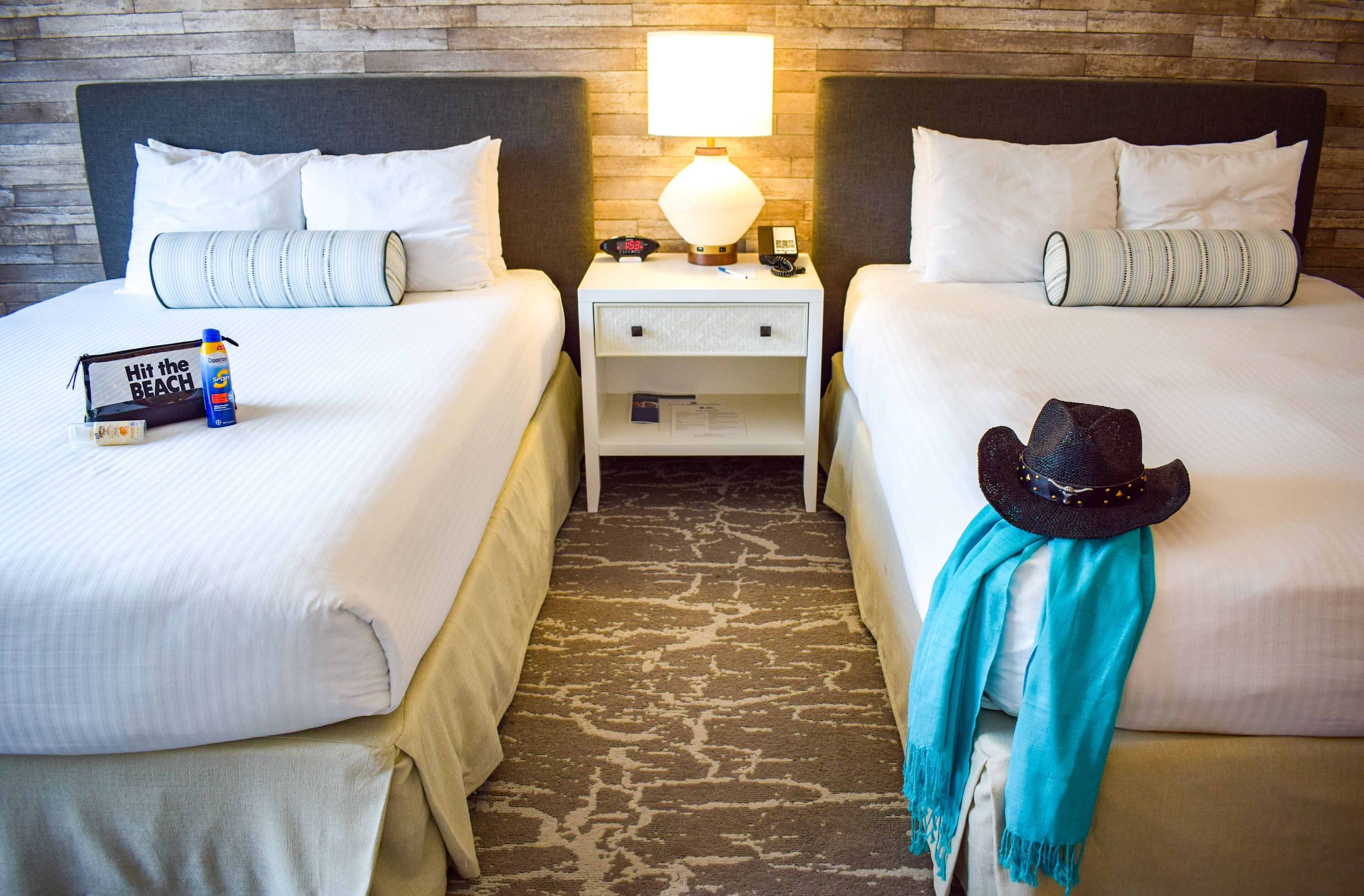 A hotel room with two beds, a nightstand with a lamp, a hat, and a scarf on one bed.