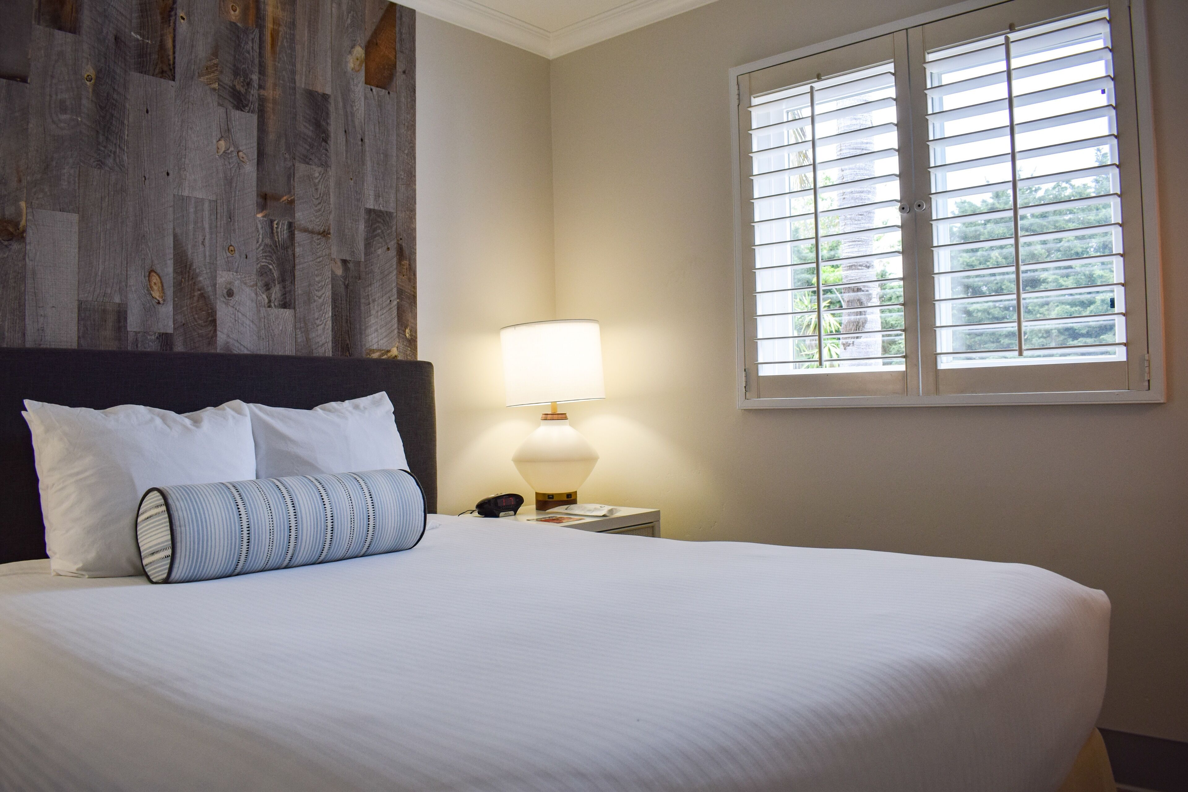 A neatly made bed with white linens and a striped pillow, a bedside lamp, and a window with blinds.