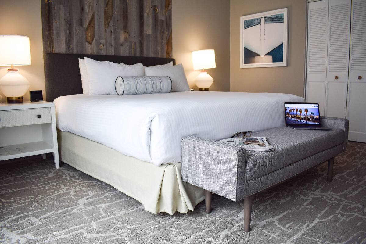 A neatly made bed with white linens, a laptop on a grey ottoman, and a magazine.