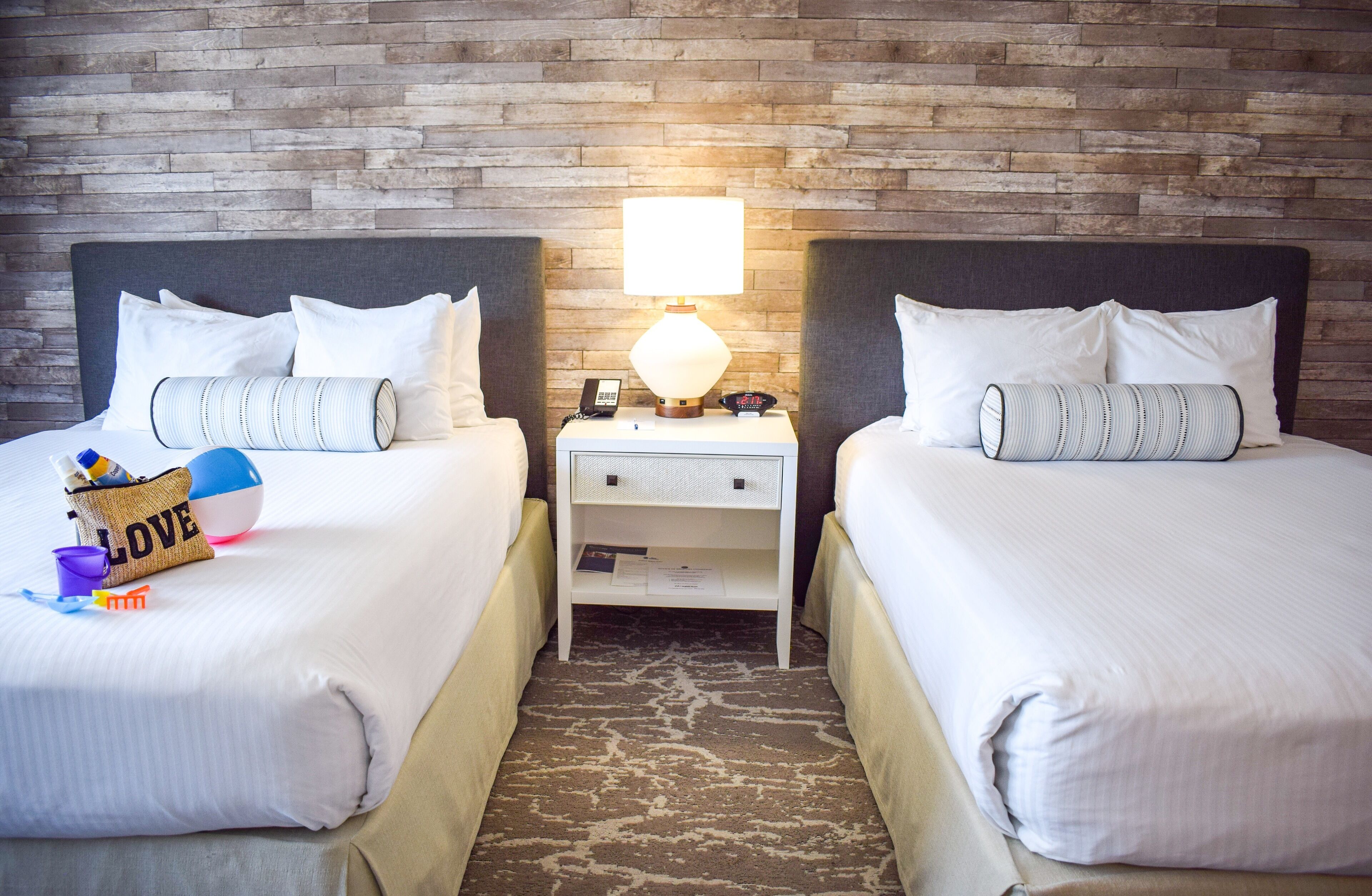 A hotel room with two beds, a nightstand with a lamp, and a brick wall.