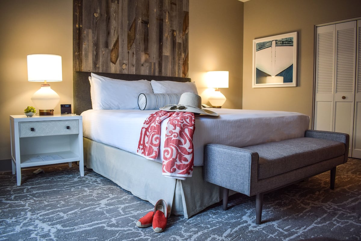 A neatly made bed with pillows and a blanket, a bedside table with a lamp, a chair, and a pair of shoes on the floor.