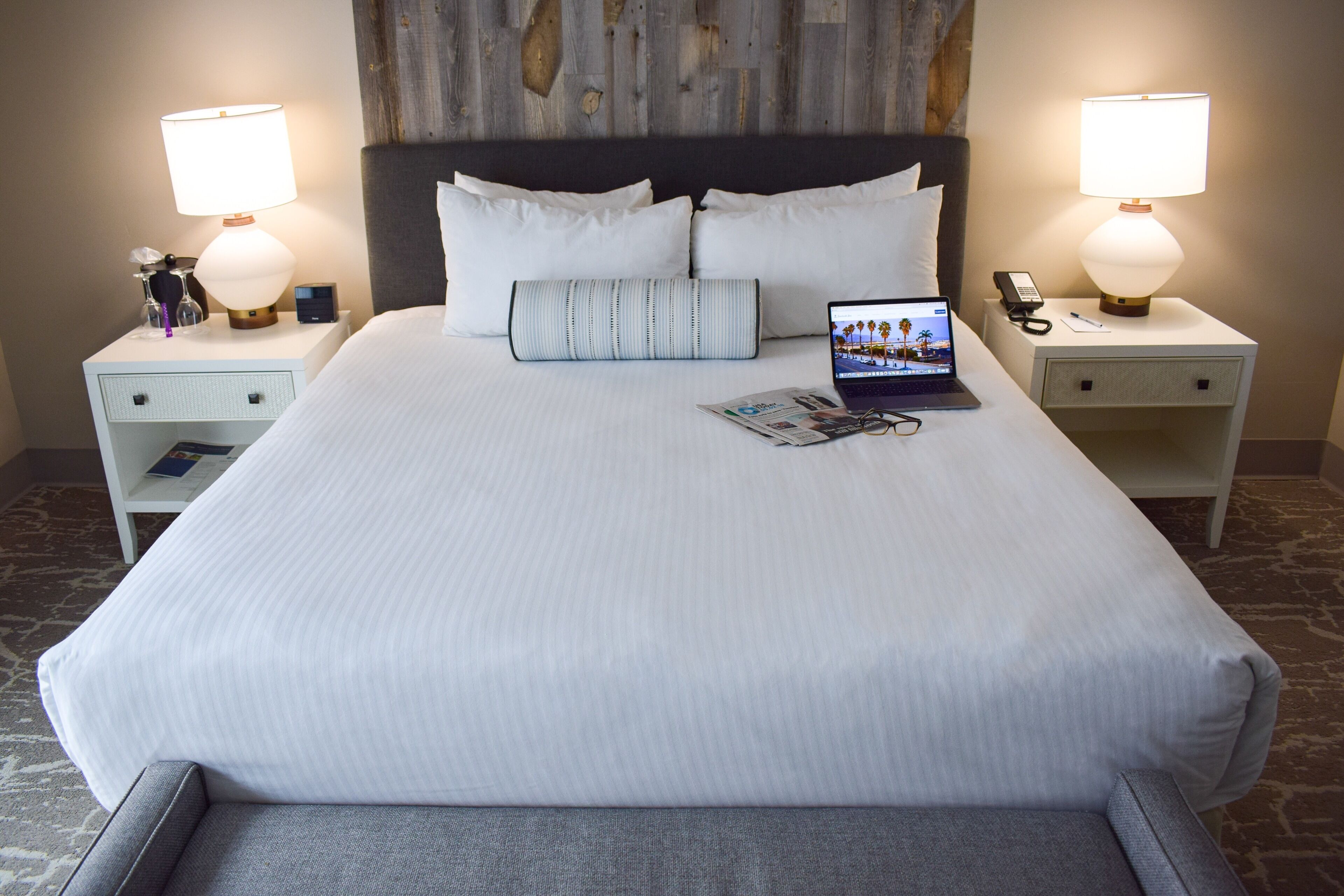 Un lit bien fait, avec du linge de lit blanc, un ordinateur portable posé sur le lit et un magazine sur la table de chevet. Il y a deux tables de chevet, chacune avec une lampe et une horloge.