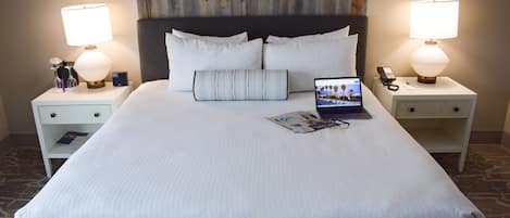 Un lit bien fait, avec du linge de lit blanc, un ordinateur portable posé sur le lit et un magazine sur la table de chevet. Il y a deux tables de chevet, chacune avec une lampe et une horloge.
