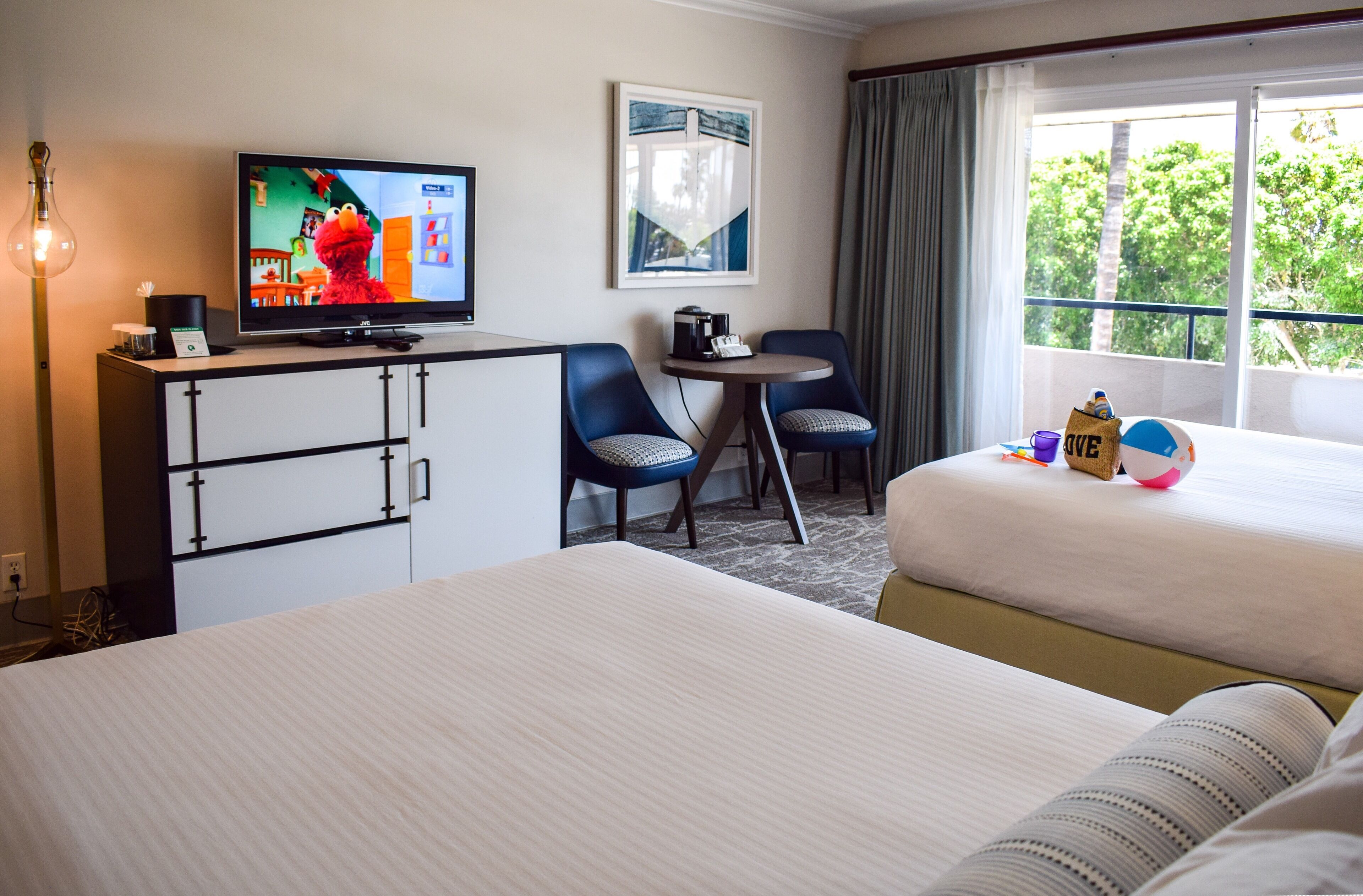 Ein Hotelzimmer mit zwei Betten, einem Fernseher, einem Schreibtisch mit Stuhl und einem Fenster mit Blick auf Bäume.