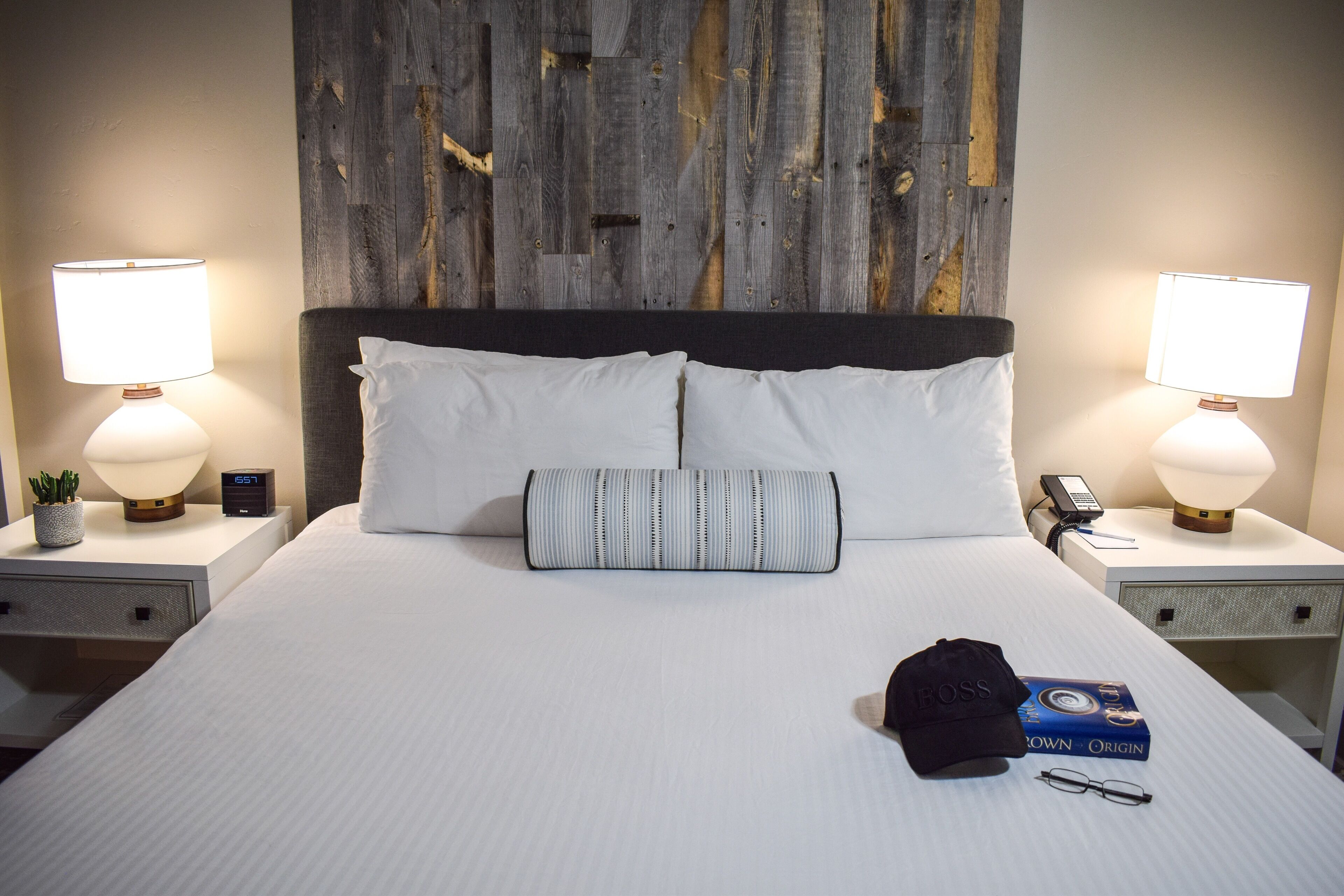 A neatly made bed with white linens, a striped pillow, a black cap, and a book on the bed. There are two bedside tables with lamps and a clock on one of them.