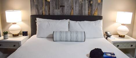 A neatly made bed with white linens, a striped pillow, a black cap, and a book on the bed. There are two bedside tables with lamps and a clock on one of them.