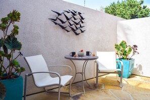 A patio with two white chairs, a small round table with two black mugs, and a decorative wall piece.