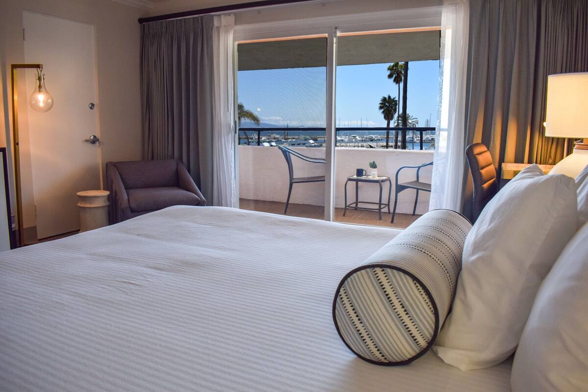 A hotel room with a large bed, a chair, a small table, and a view of the beach and palm trees through the balcony doors.