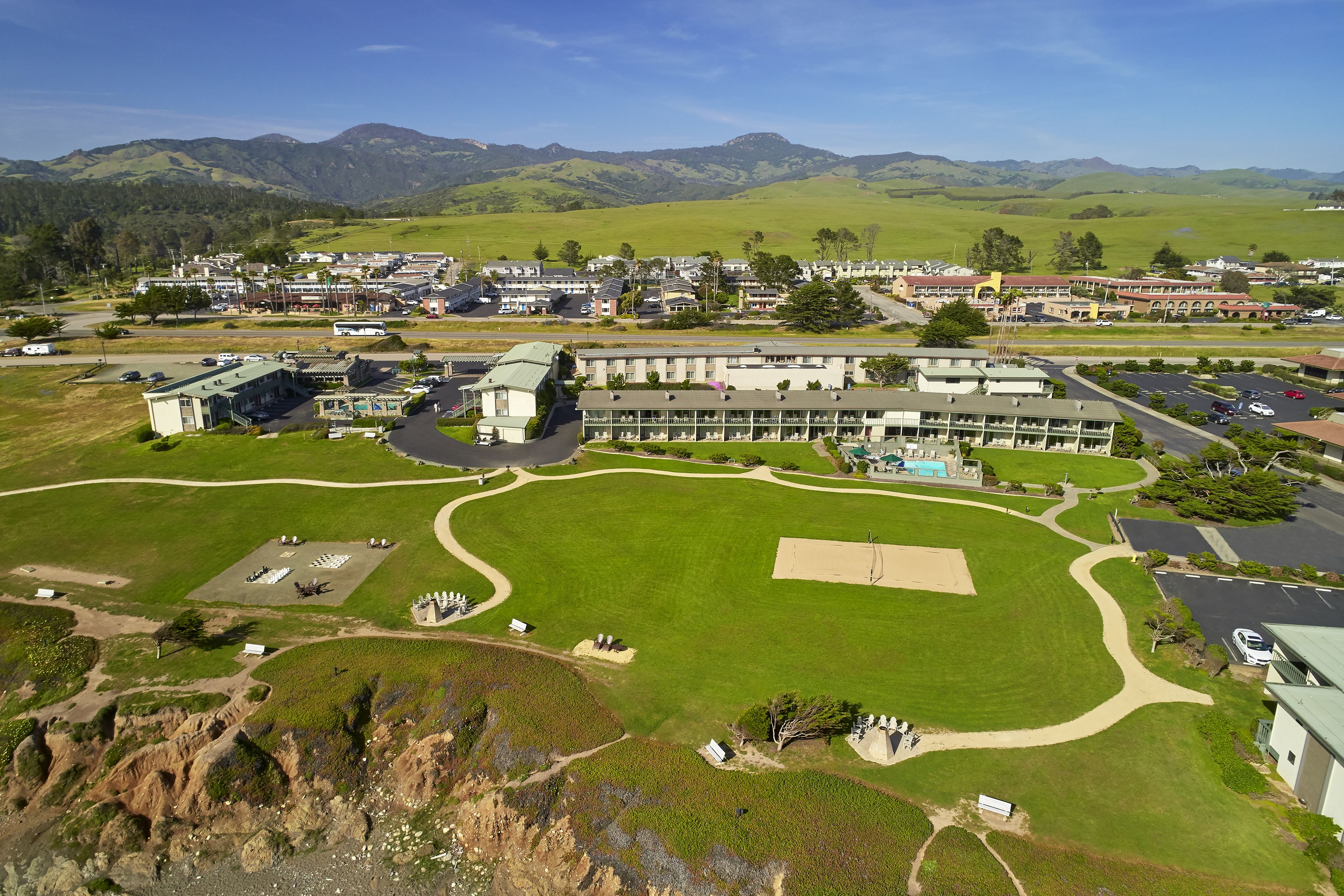 A large building with multiple wings, surrounded by green fields and mountains.