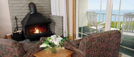 A living room with a fireplace, a sofa, a small table with a vase of flowers, and a view of the ocean through large windows.