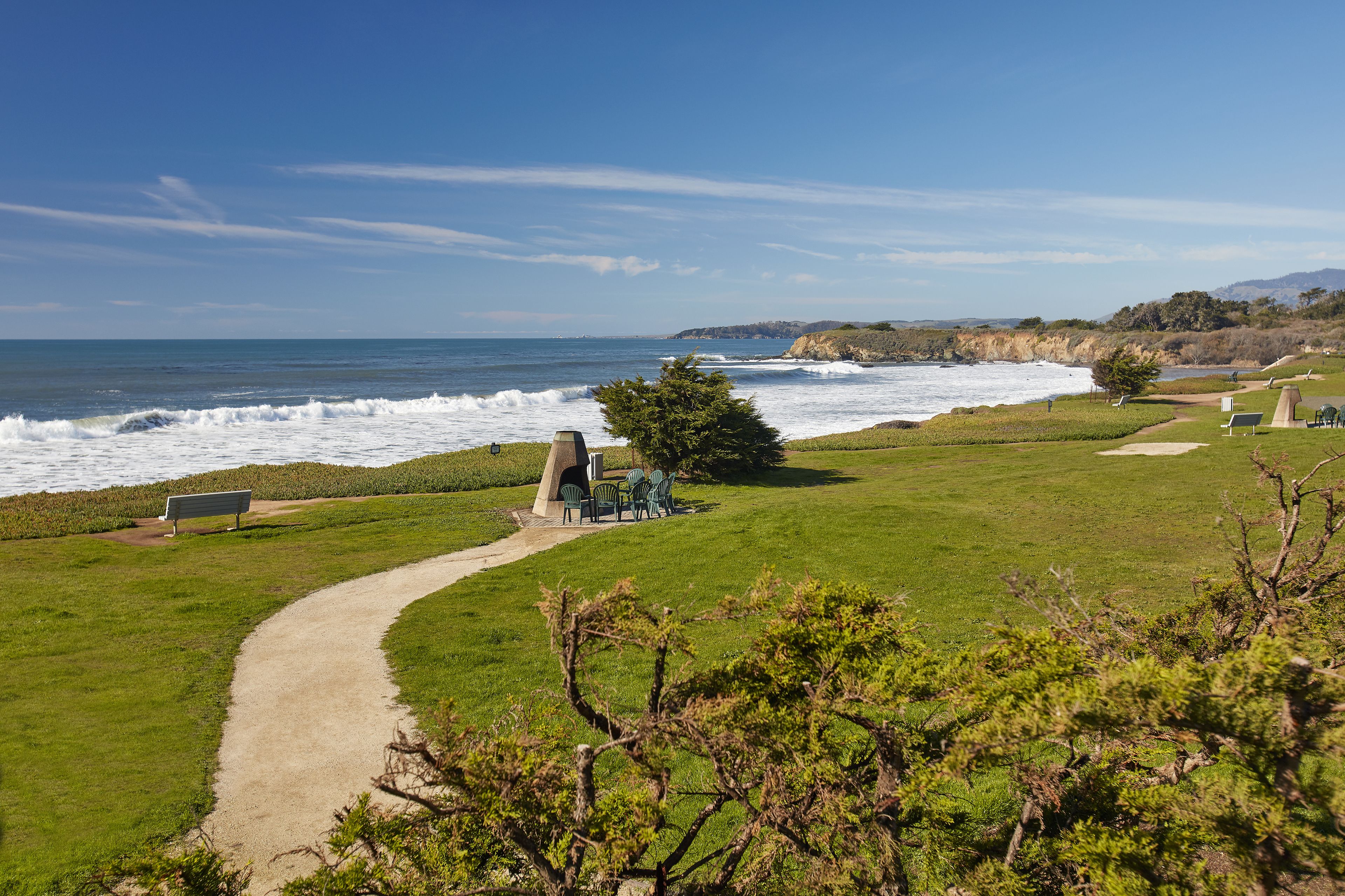 A coastal park with a paved path, benches, and green chairs overlooking the ocean.