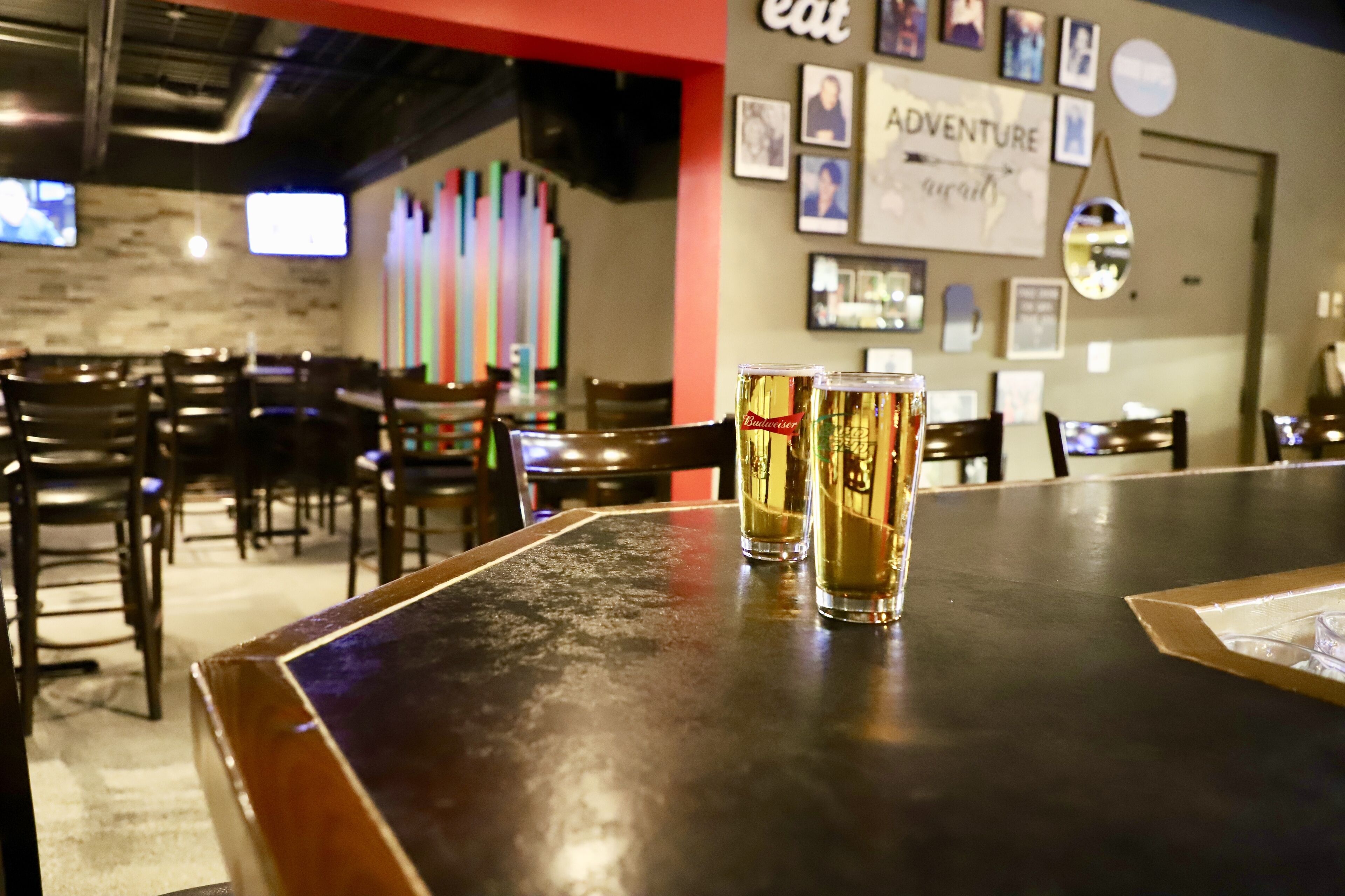 A bar area with two glasses of beer on a table, surrounded by chairs and a wall with framed pictures and the word "ADVENTURE".