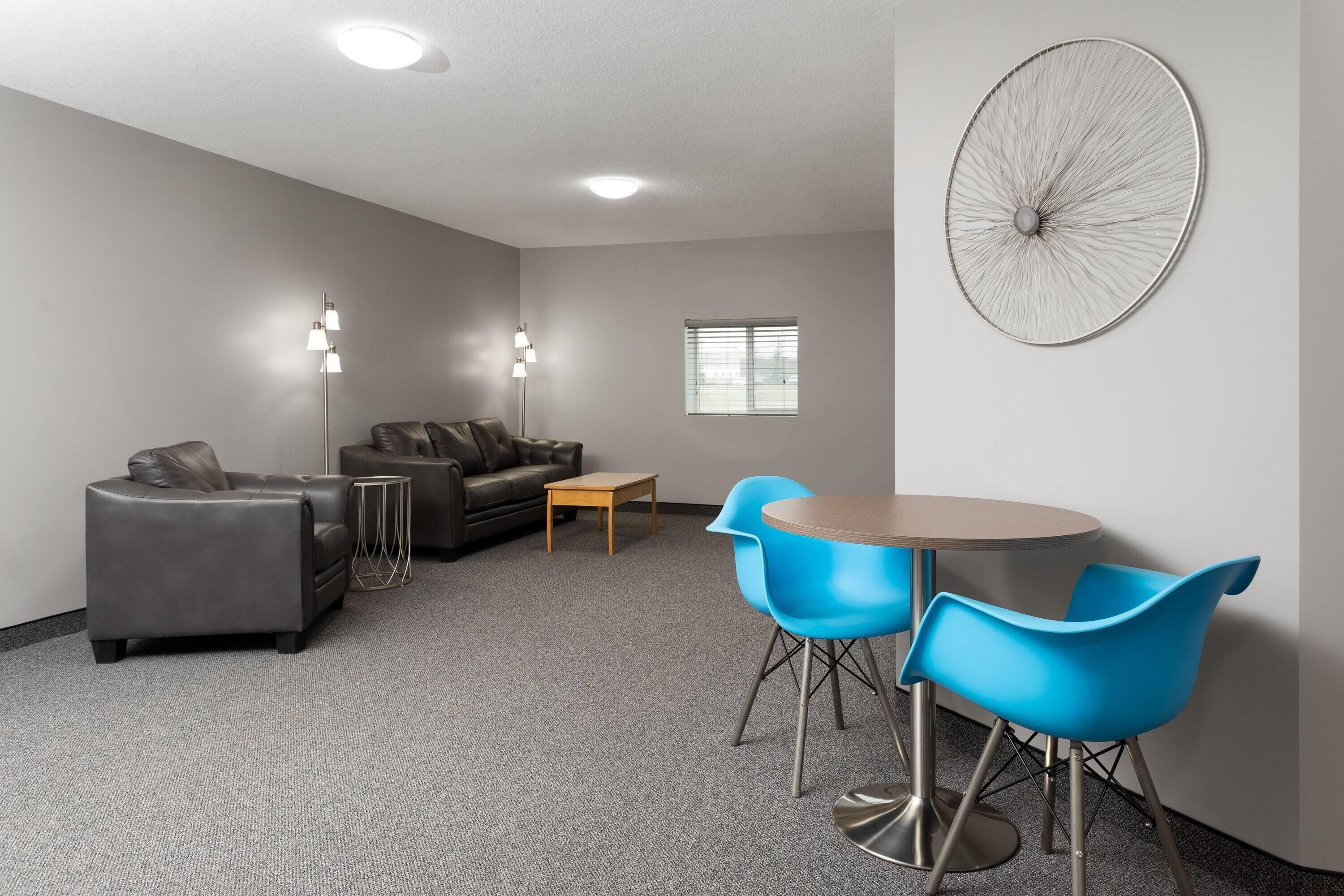 A modern living room with a gray sofa, a coffee table, and blue chairs. There is a large wall art piece and a window with blinds.