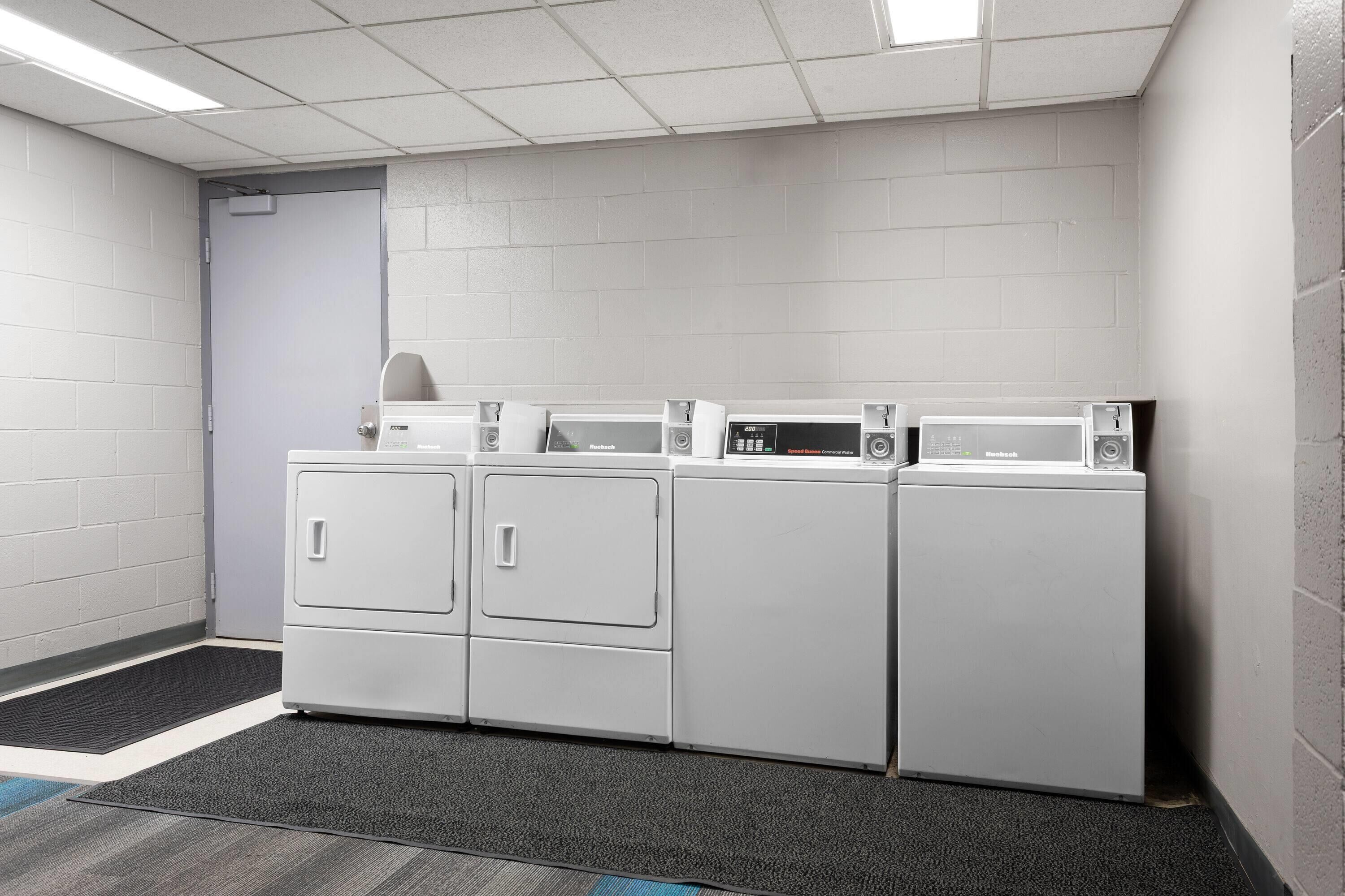 Une buanderie équipée de quatre machines à laver et de quatre sèche-linge à chargement frontal, tous de couleur blanche, ainsi que d'un tapis gris et d'un mur recouvert de carreaux blancs.