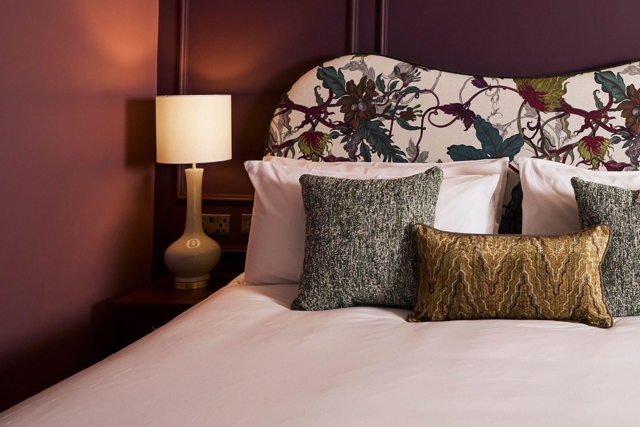 Una cama impecablemente arreglada con sábanas blancas y tres almohadones decorativos de distintos diseños. Se ve una mesita de noche con una lámpara y un pequeño adorno.