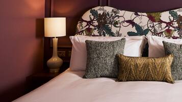 A neatly made bed with white linens and three decorative pillows in different patterns. A bedside table with a lamp and a small decorative item is visible.