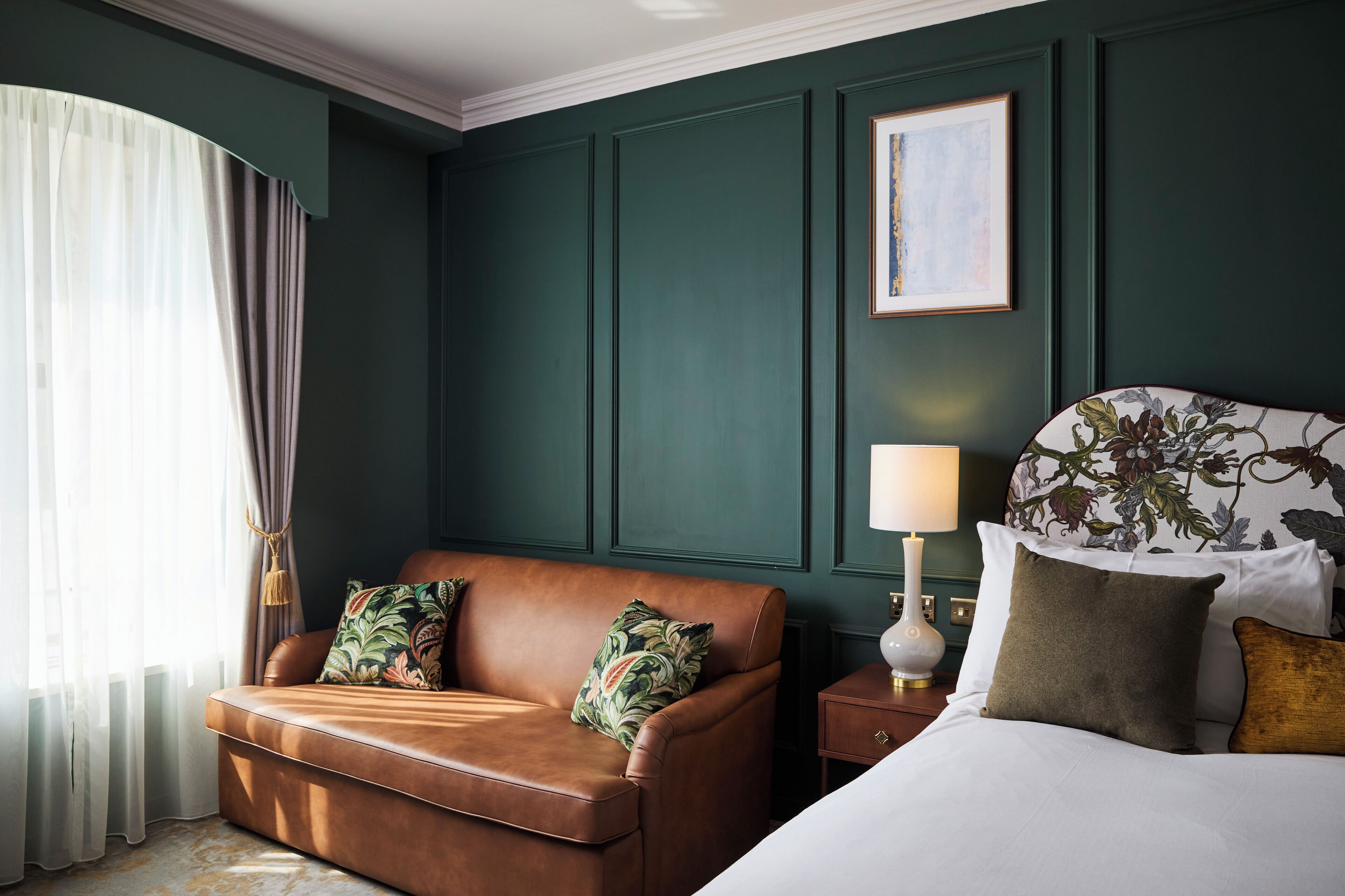 A room with a brown leather sofa, a bed with white bedding, a nightstand with a lamp, and a framed picture on the wall.