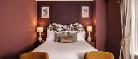 A hotel room with a large bed, two bedside lamps, a decorative headboard, and two armchairs.