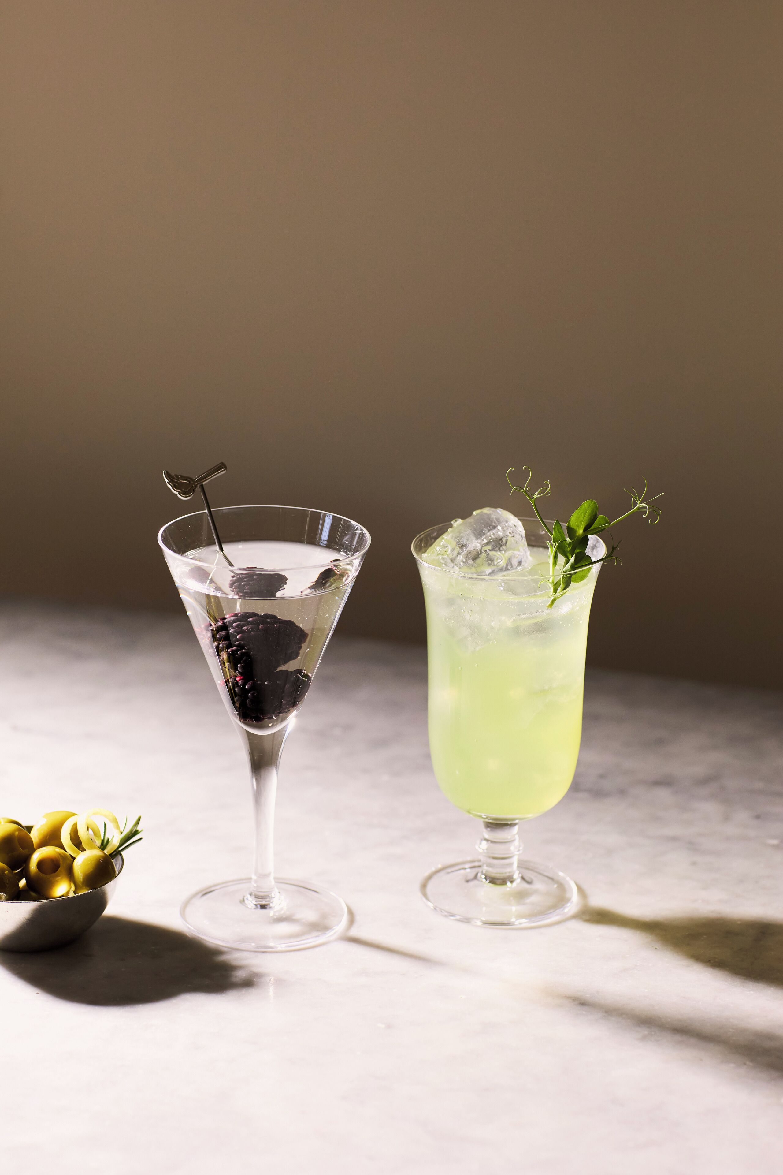 Two drinks in martini glasses, one with a blackberry and the other with a green drink and a sprig of herb.