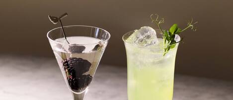 Two drinks in martini glasses, one with a blackberry and the other with a green drink and a sprig of herb.