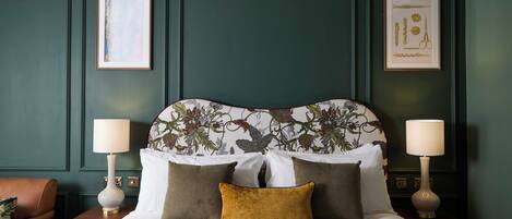 Una cama bien hecha con un cabecero floral, dos lámparas de noche y un sillón marrón.