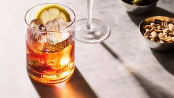 Two glasses of beverages on a marble surface, one with a lemon slice and the other with a cocktail stirrer.