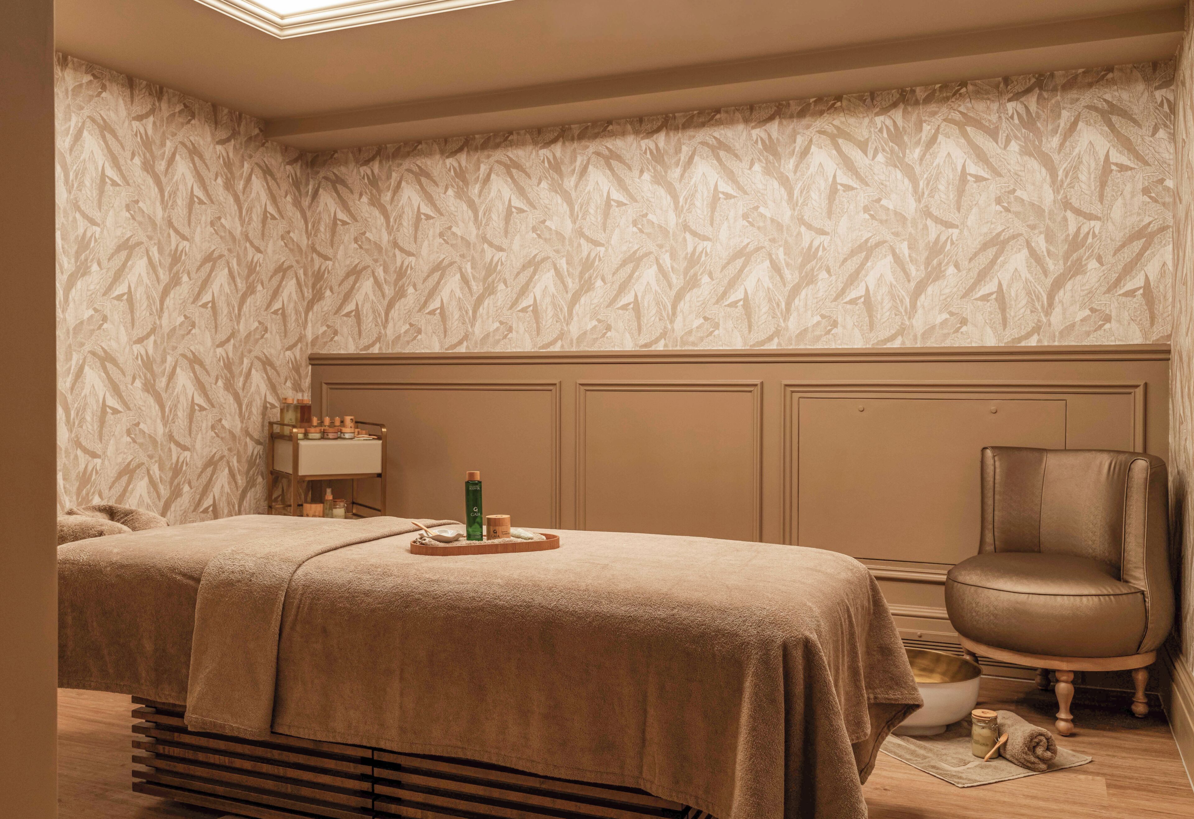 Un cuarto de spa con una mesa de masaje, una silla, una mesita con botellas y un cuenco decorativo.