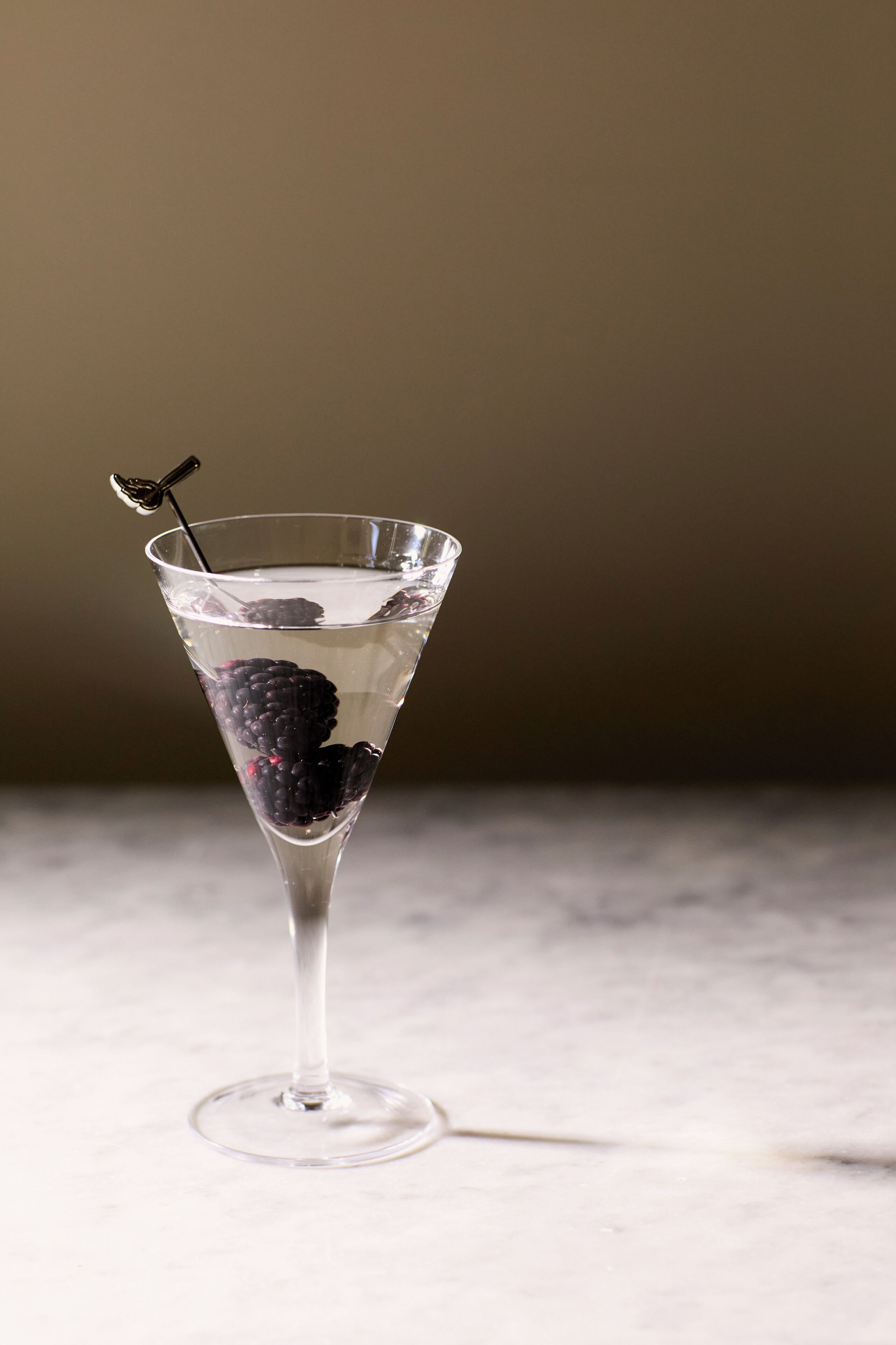 Una copa de martini con zarzamoras y un removedor para cócteles sobre una superficie de mármol.