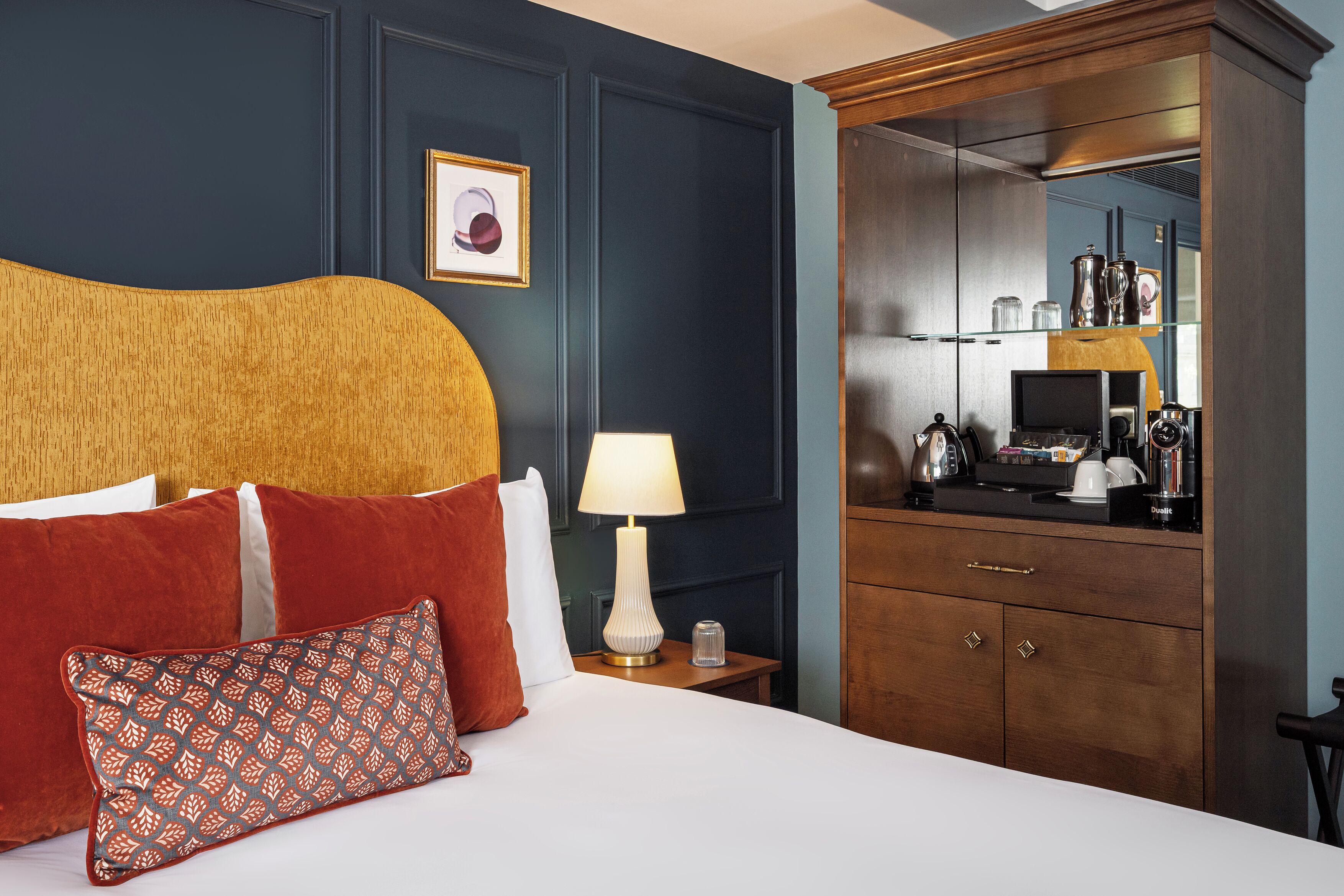 Quarto de hotel com uma cama grande, uma mesa de cabeceira com abajur, penteadeira de madeira e um espelho refletindo o quarto.