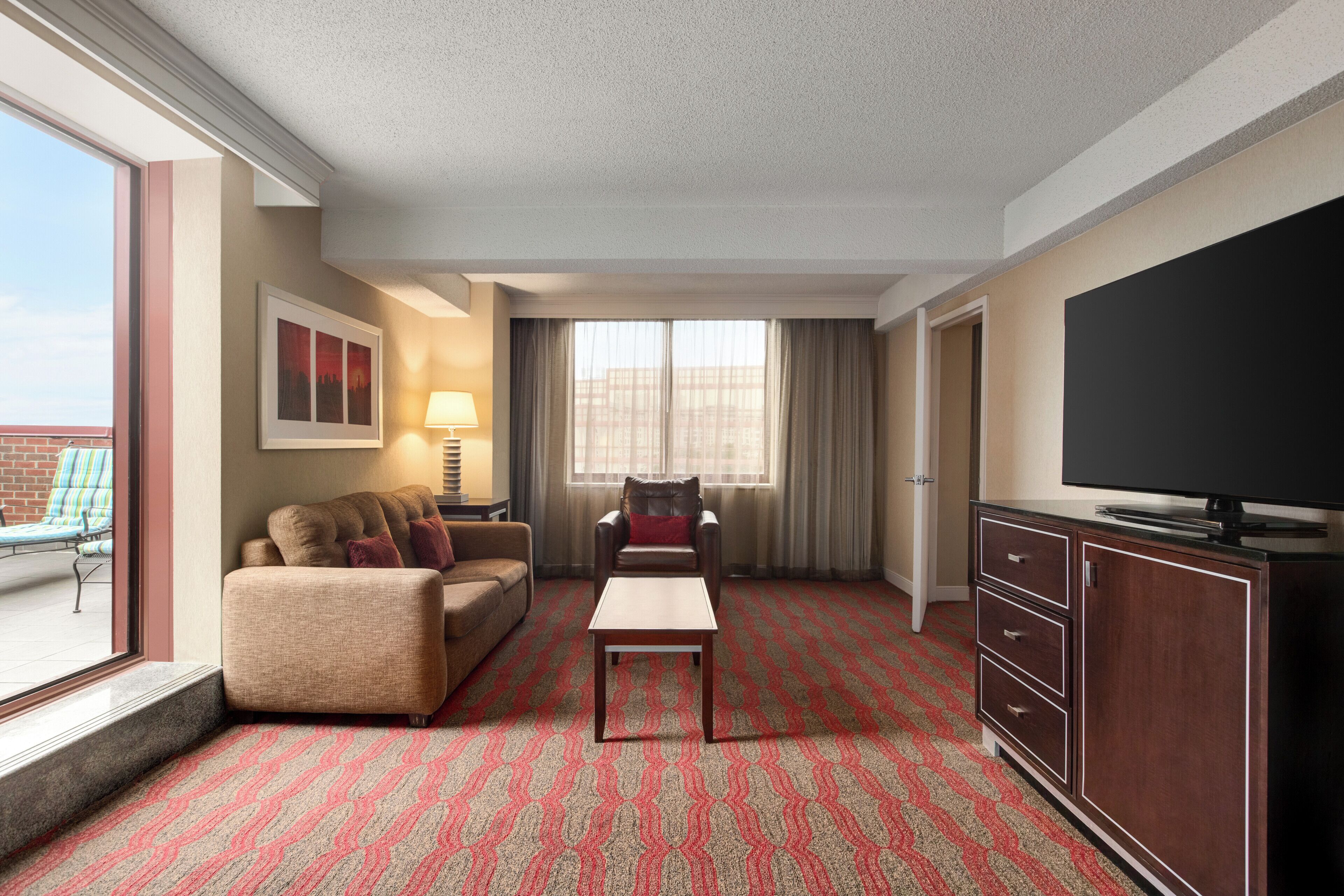 A hotel room with a sofa, armchair, TV, and a patterned carpet.