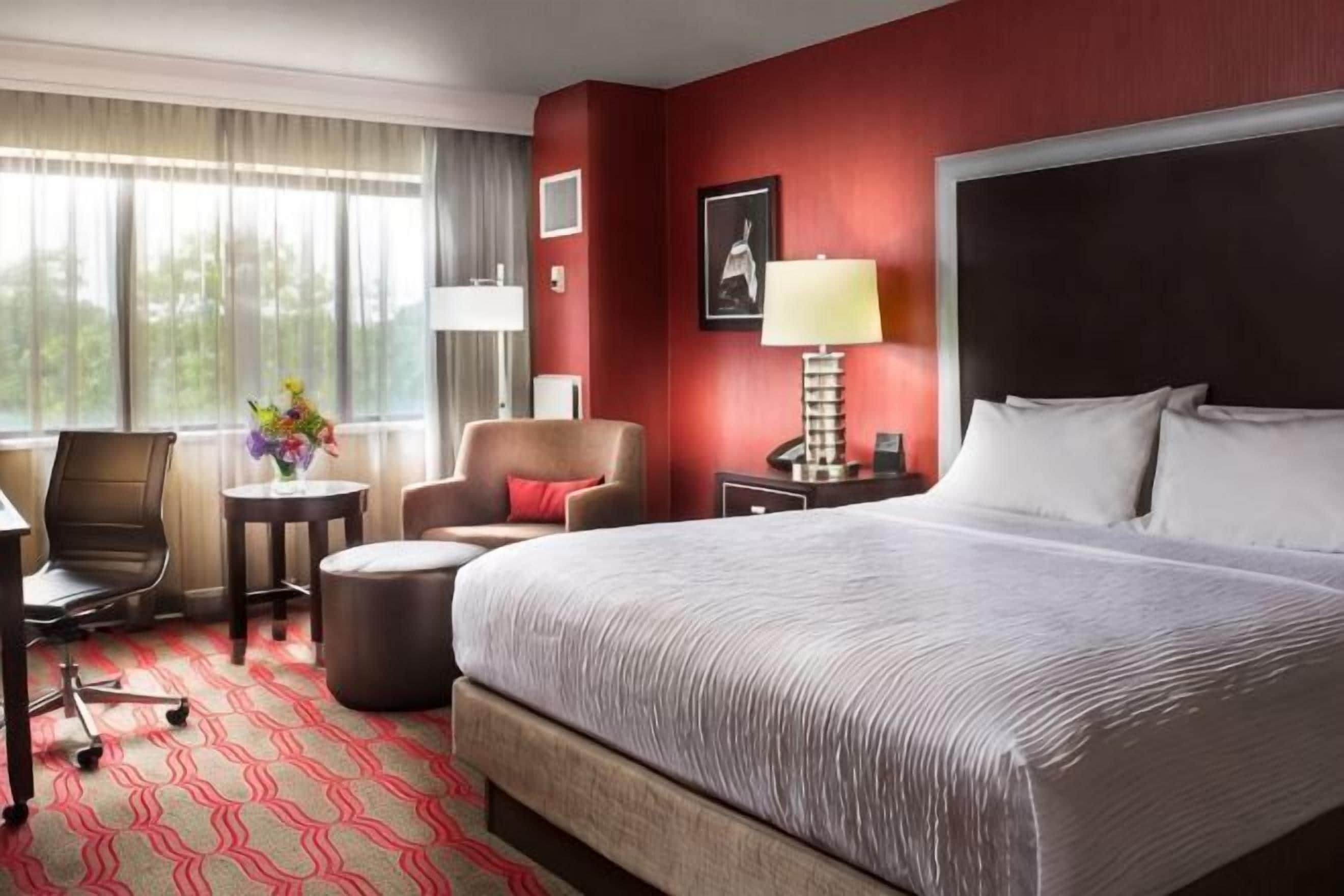 A hotel room with a large bed, a desk, a chair, a small table, and a window with curtains.