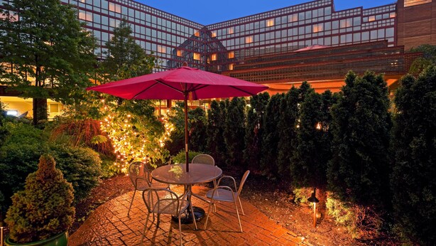 An outdoor patio with a table and chairs, surrounded by trees and a building with large windows in the background. - DoubleTree by Hilton New York LaGuardia Airport (East Elmhurst)