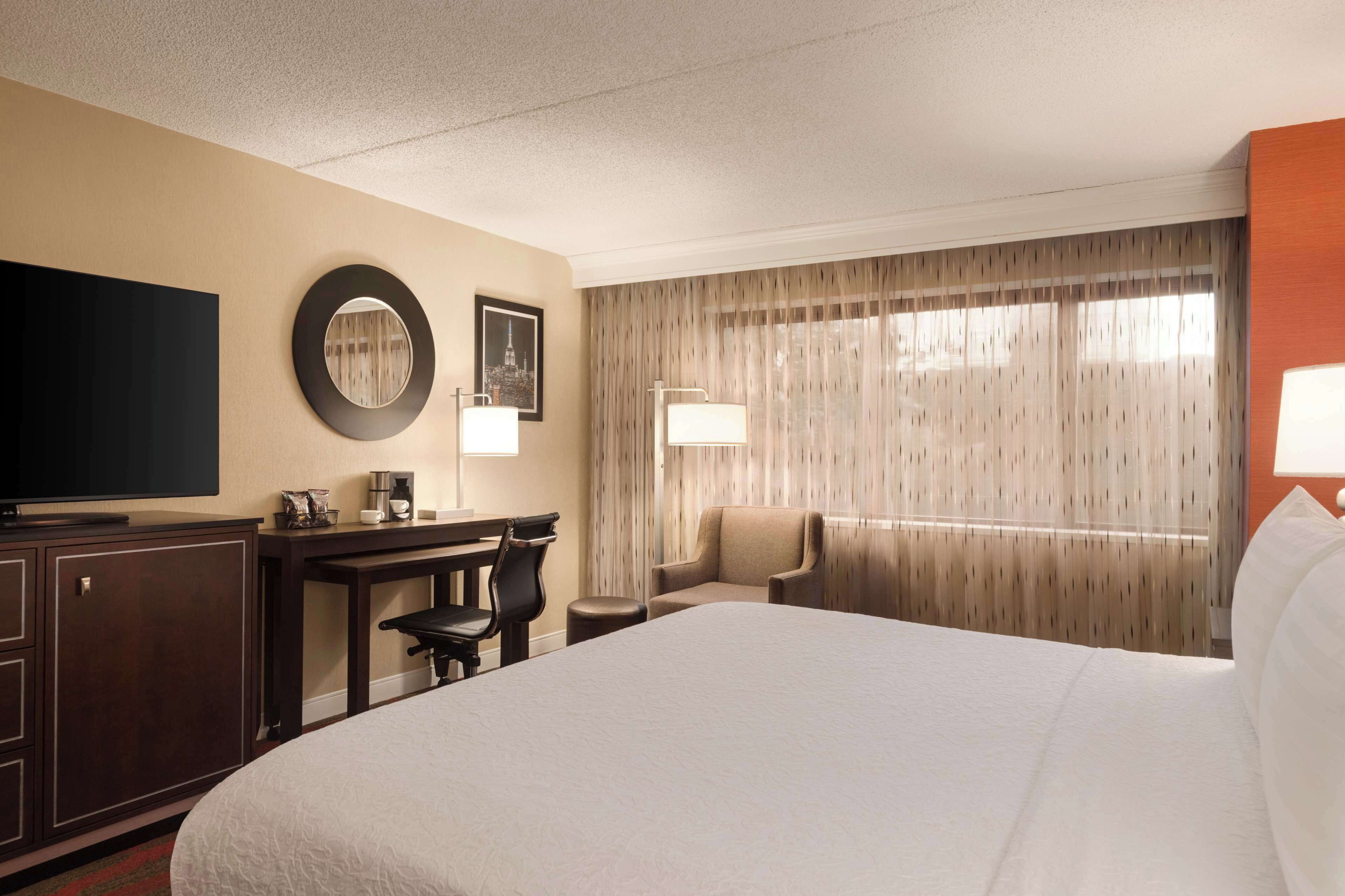Ein Hotelzimmer mit einem großen Bett, einem Schreibtisch mit Stuhl, einem Fernseher, einer Kaffeemaschine und einem Fenster mit Vorhängen.