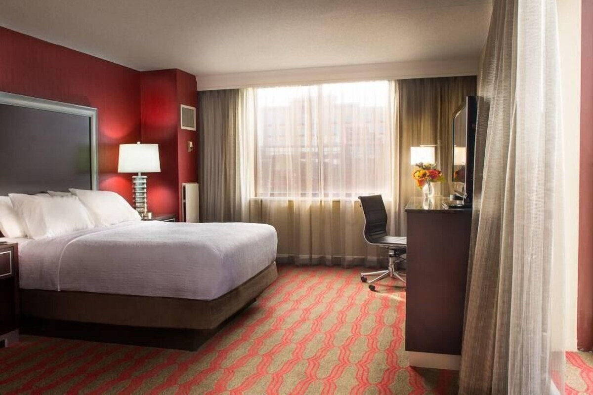 A hotel room with a bed, a desk, a chair, a TV, and a window with curtains.