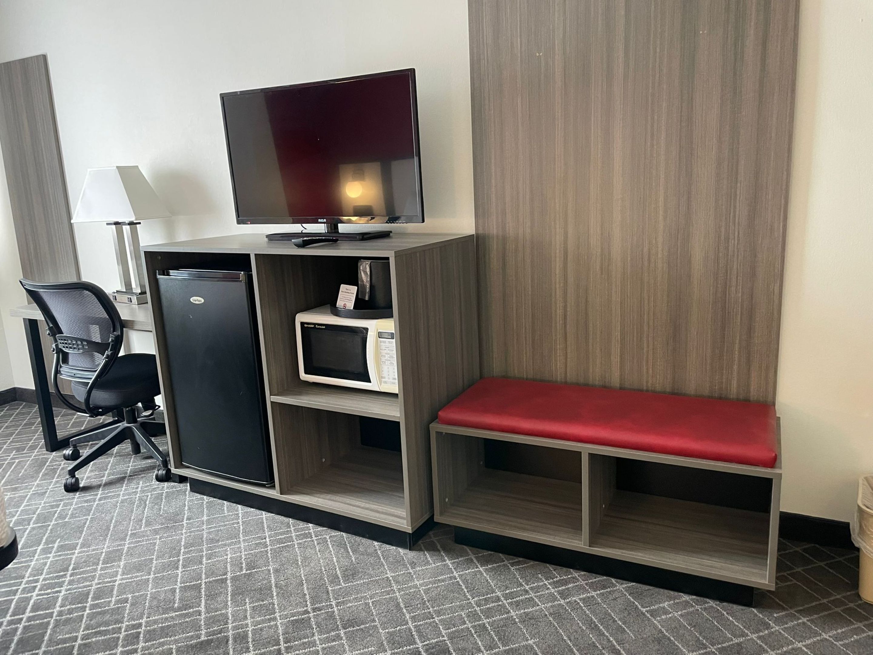 A compact hotel room with a kitchenette, a flat-screen TV, a red bench, and a desk with a lamp.
