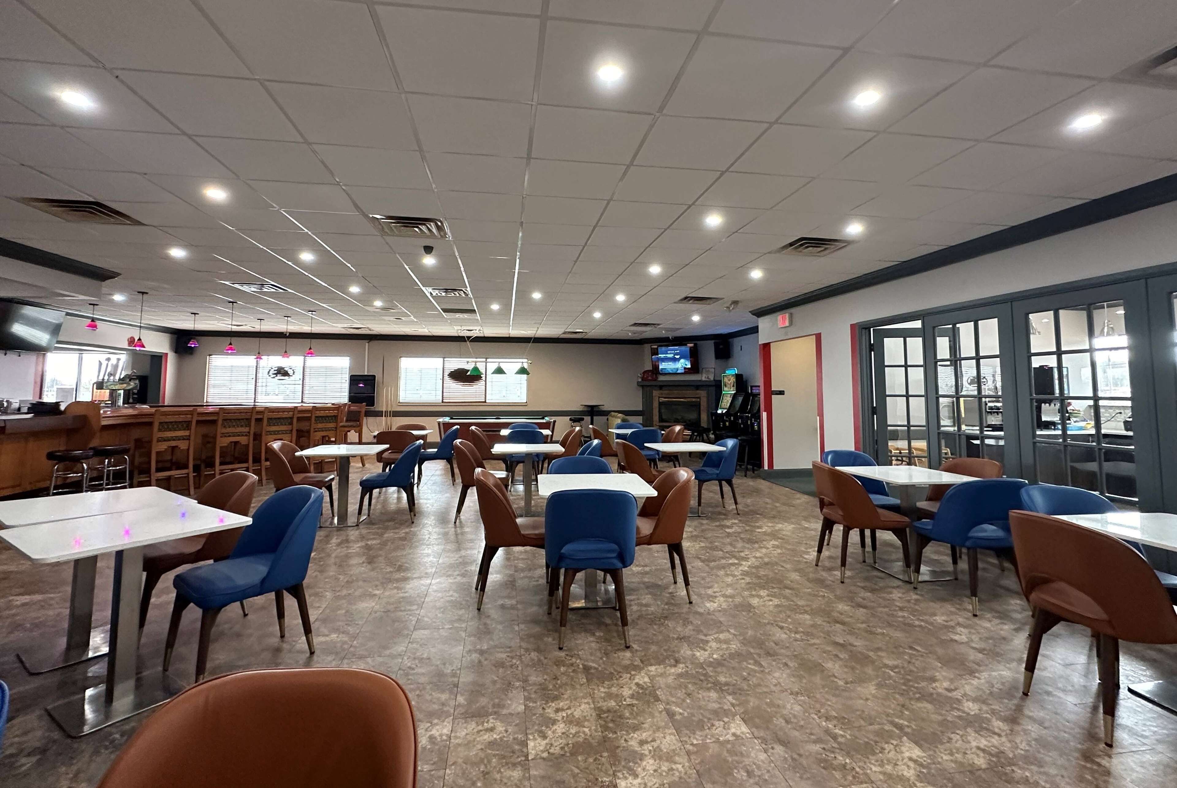 A spacious cafeteria with multiple tables and chairs, a counter area, and large windows.